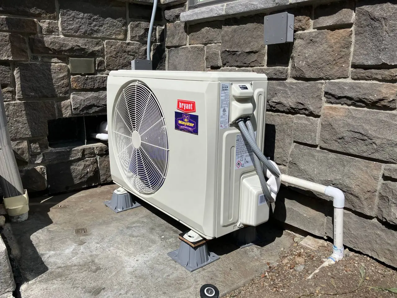 White Bryant heat pump unit installed on riser feet against a stone exterior wall.
