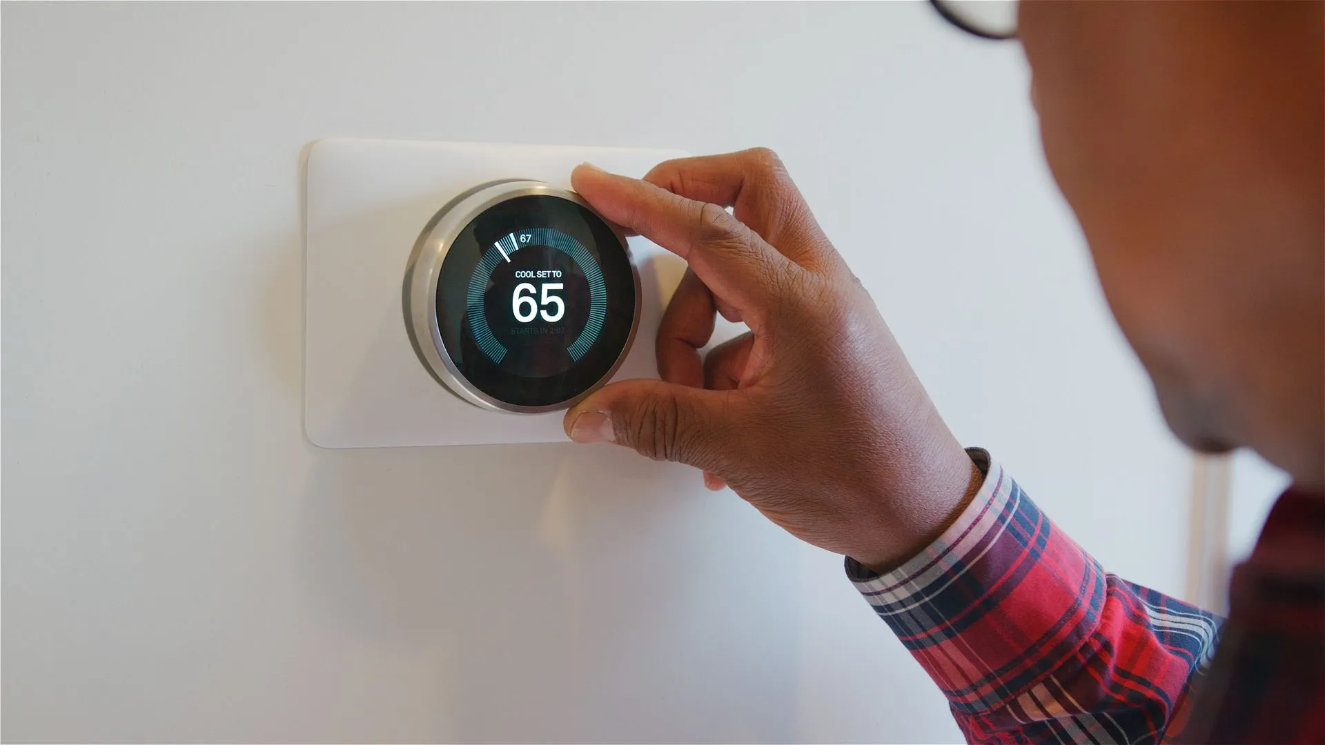 Hand turning the dial on a round smart thermostat mounted on a white wall to set cooling temperature.