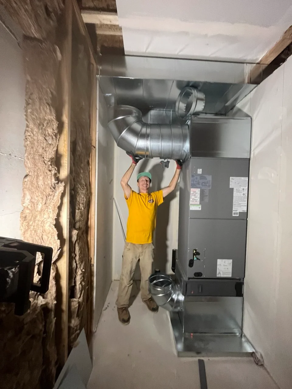 A man installs shiny metal ducts on a gray furnace unit, showing local Heating System Services Greater Puget Sound.