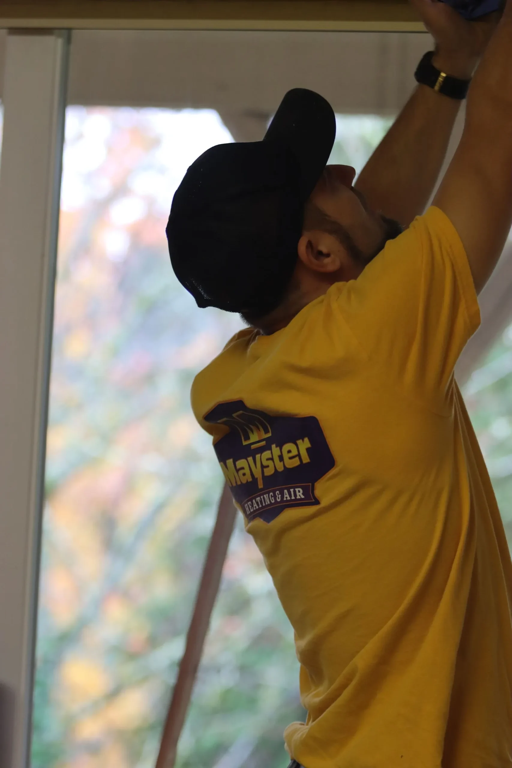 Technician in yellow Mayster Heating and Air shirt reaching up to work on an overhead indoor HVAC unit.