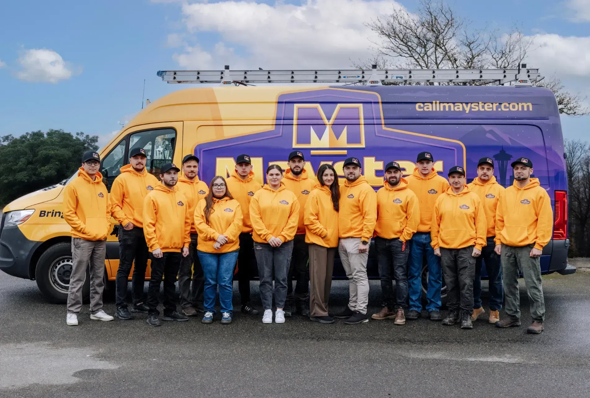 Group of HVAC technicians and staff in yellow hoodies standing together in front of a Mayster service van outdoors.