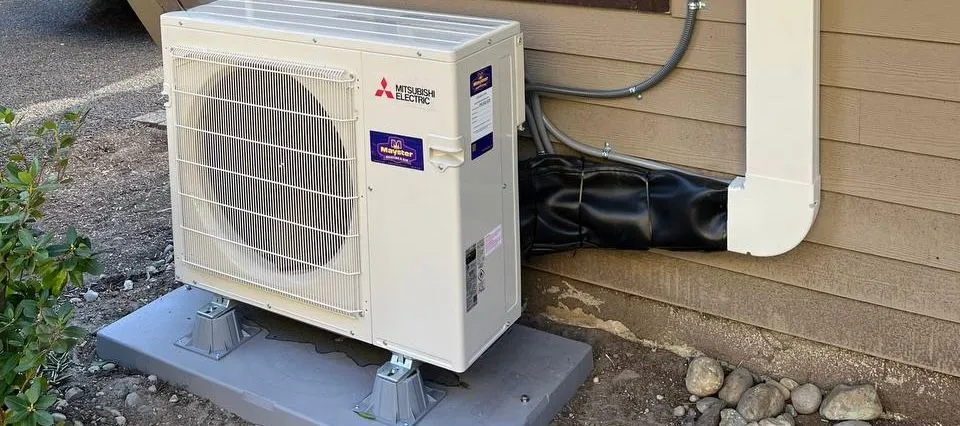 White Mitsubishi Electric outdoor heat pump unit mounted on a gray base next to an exterior wall.